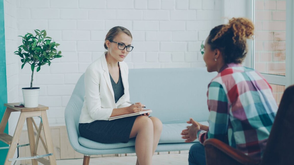 Therapist in a white blazer and glasses takes notes as she listens to a client in a bright, modern therapy room.