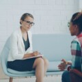 Therapist in a white blazer and glasses takes notes as she listens to a client in a bright, modern therapy room.