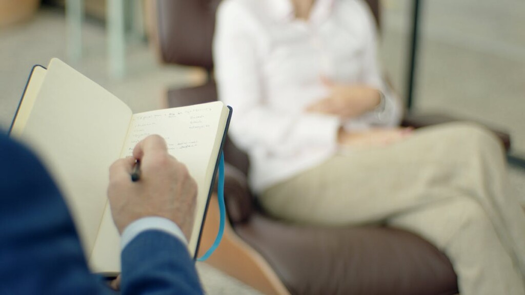 Person writes in an open notebook held on their lap in a cozy lounge with a blurred figure seated in the background.