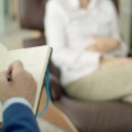 Person writes in an open notebook held on their lap in a cozy lounge with a blurred figure seated in the background.