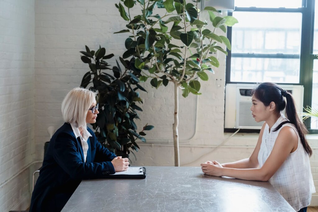 Two women sit across from each other at a metal table in a bright office; one with a clipboard, the other listening attentively.
