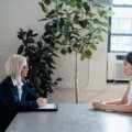 Two women sit across from each other at a metal table in a bright office; one with a clipboard, the other listening attentively.