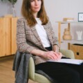 Woman with brown hair sits in a light-green chair, writing in a notebook while wearing a plaid blazer in a bright office.