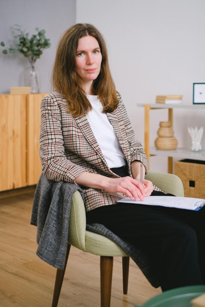 Woman with brown hair sits in a light-green chair, writing in a notebook while wearing a plaid blazer in a bright office.
