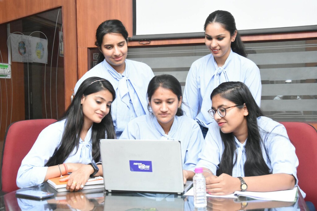 Five young women in light blue blouses gather around a laptop on a glass table in a meeting room.