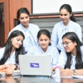 Five students in light blue blouses gather around a laptop, collaborating on a project at a conference table in a classroom setting.